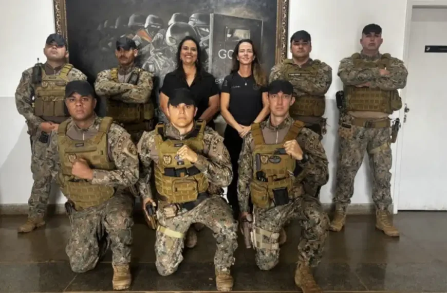 PPGO tem novo uniforme-Um grupo de policiais penais aparece em formação para foto oficial. Eles vestem uniformes camuflados novos e coletes táticos padronizados. Atrás, duas mulheres em trajes civis posam ao centro, enquanto ao fundo há um quadro com imagem de operações policiais.