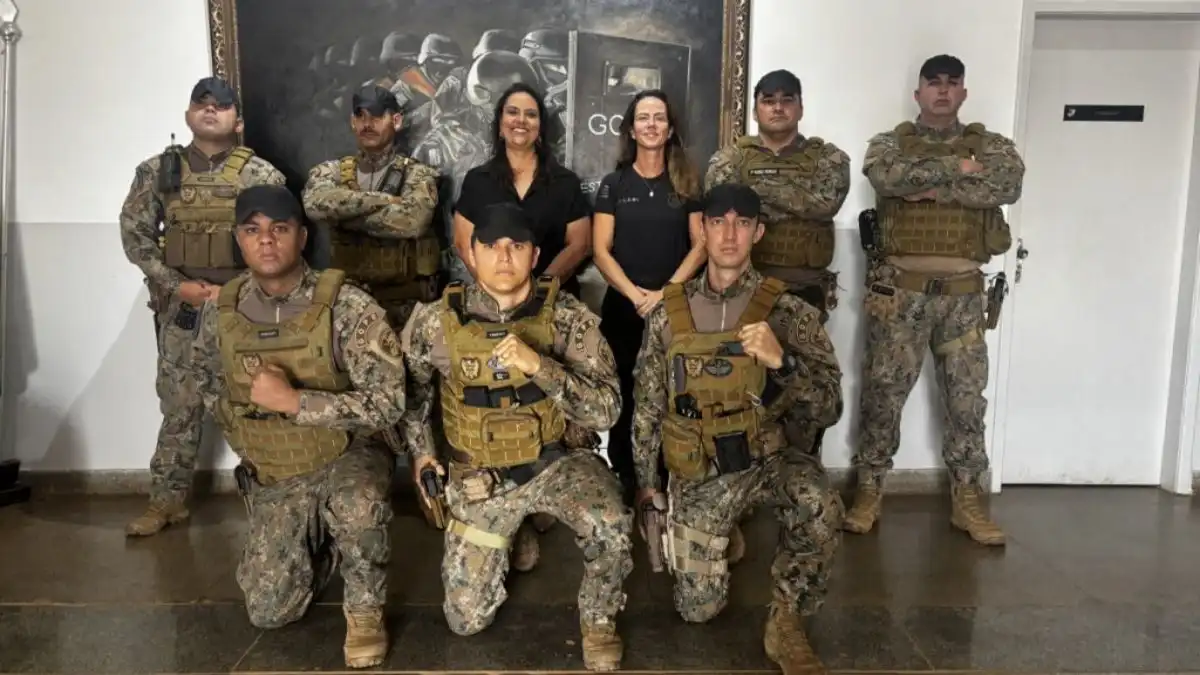 PPGO tem novo uniforme-Um grupo de policiais penais aparece em formação para foto oficial. Eles vestem uniformes camuflados novos e coletes táticos padronizados. Atrás, duas mulheres em trajes civis posam ao centro, enquanto ao fundo há um quadro com imagem de operações policiais.