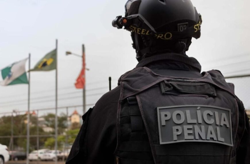 Homem fingia ser Policial Penal - Agente da Polícia Penal, de uniforme e colete tático, aparece de costas com a inscrição ‘Polícia Penal’, diante de bandeiras hasteadas ao fundo