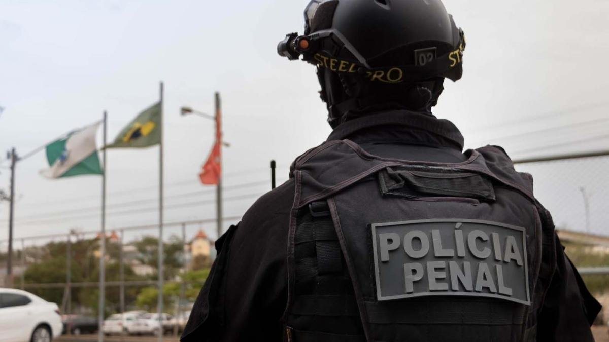 Homem fingia ser Policial Penal - Agente da Polícia Penal, de uniforme e colete tático, aparece de costas com a inscrição ‘Polícia Penal’, diante de bandeiras hasteadas ao fundo