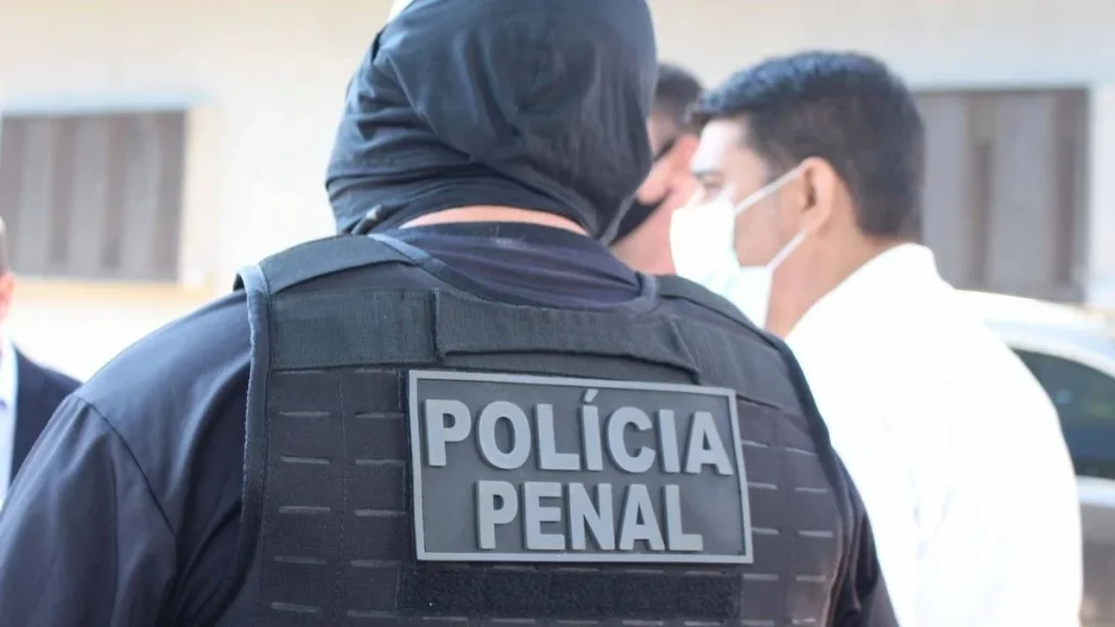 Agente de segurança com colete preto identificado como “Polícia Penal” visto de costas, usando capuz, em ambiente externo com outras pessoas ao fundo.
