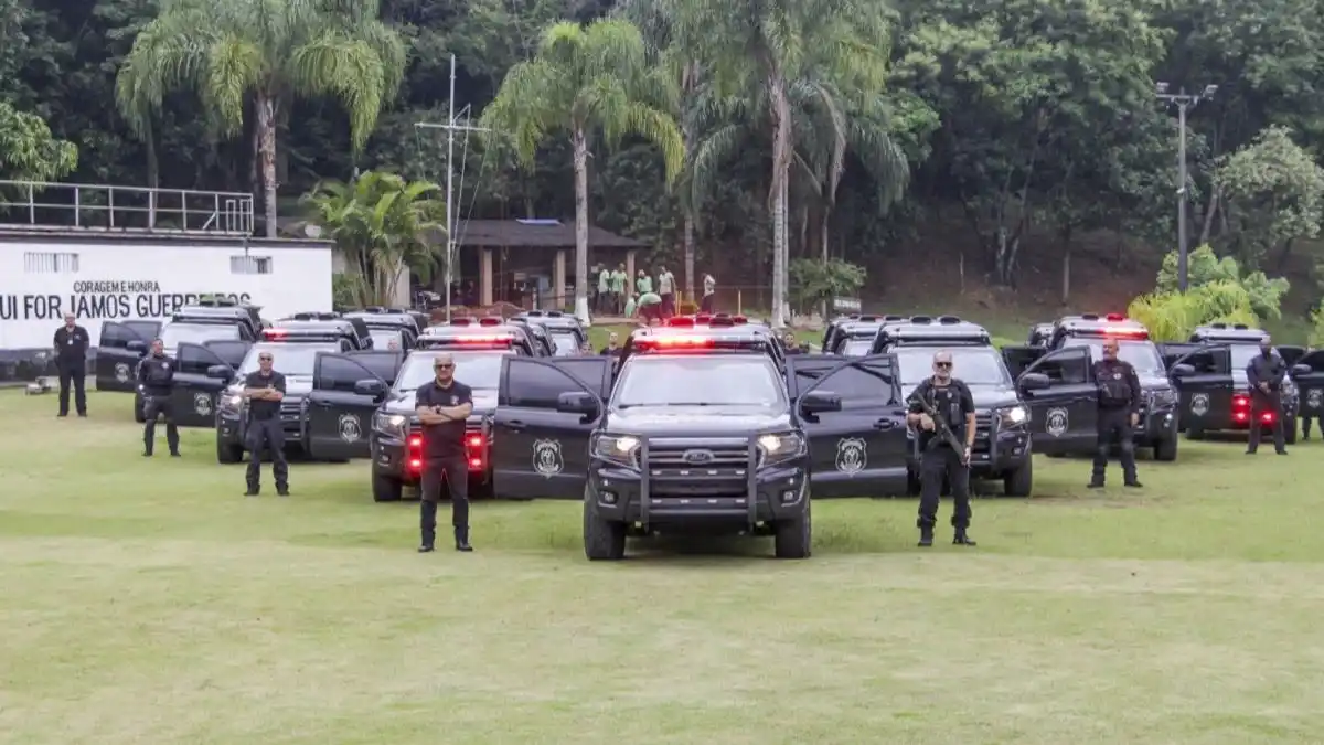 Reestruturação da PPRJ não avança-Policiais penais aparecem perfilados ao lado de viaturas pretas com sirenes ligadas, em área gramada com árvores ao fundo, em formação de exibição operacional.