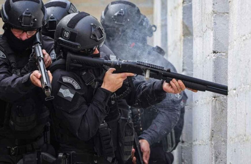 Policiais do batalhão de elite, fortemente equipados e armados, realizam ação tática junto a uma parede de concreto, com um deles efetuando disparo de arma de fogo.