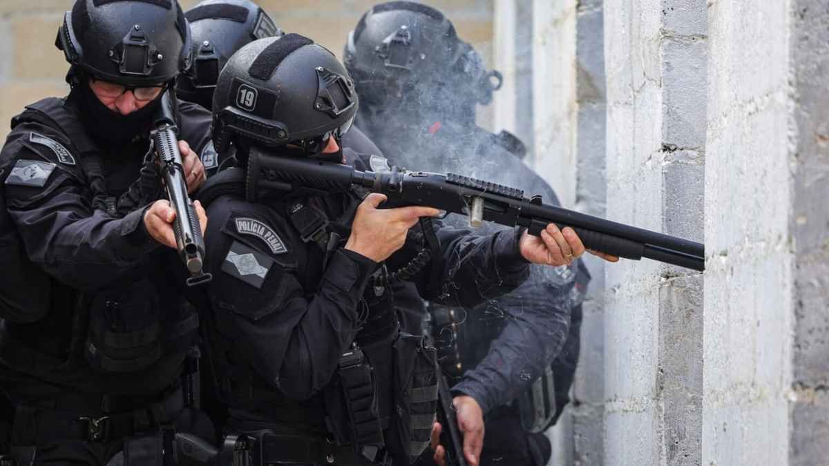 Policiais do batalhão de elite, fortemente equipados e armados, realizam ação tática junto a uma parede de concreto, com um deles efetuando disparo de arma de fogo.