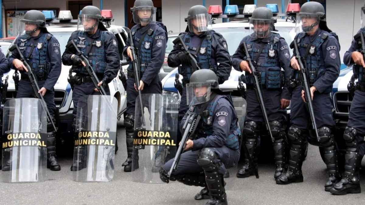 formação de choque, equipados com escudos, capacetes e armas não letais, prontos para atuação em controle de distúrbios civis.