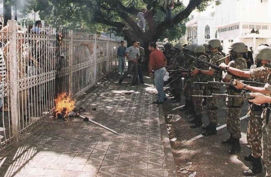 Grupo de militares armados com fuzis e escudos em formação, confrontando manifestantes atrás de um gradil. No chão, próximo à grade, há um objeto em chamas.