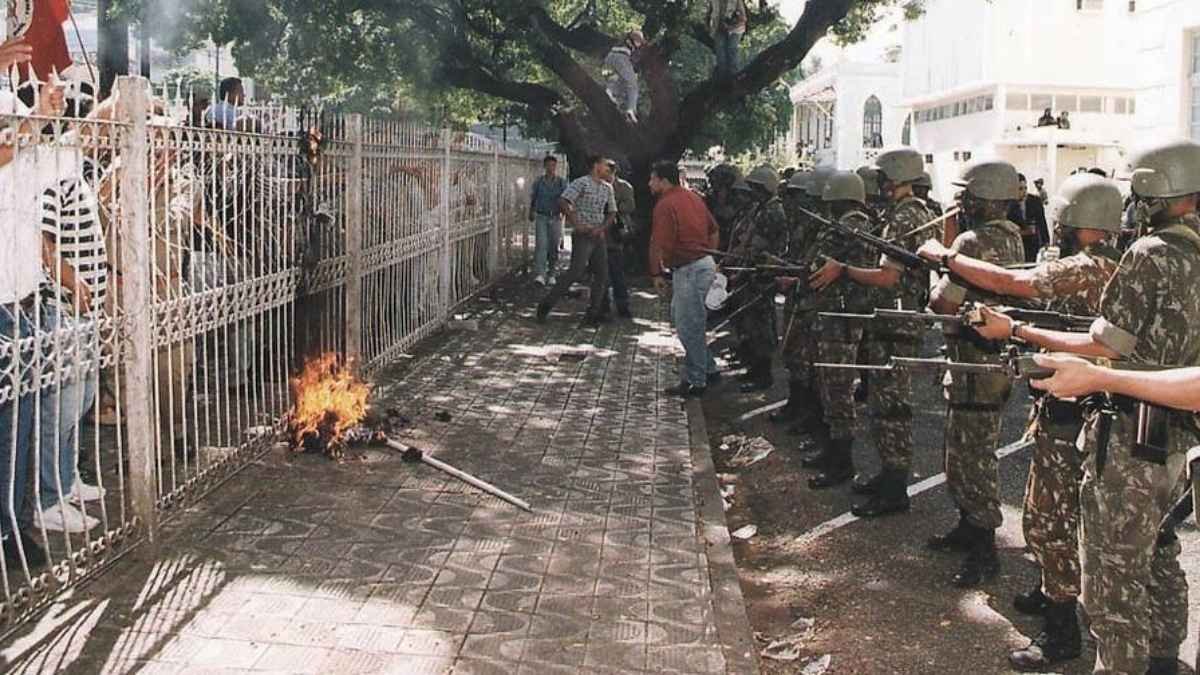Grupo de militares armados com fuzis e escudos em formação, confrontando manifestantes atrás de um gradil. No chão, próximo à grade, há um objeto em chamas.