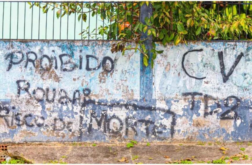 25% dos brasileiros vivem sob jugo de facções-Muro desgastado com pichações em tinta preta, incluindo mensagens de proibição e siglas de facção criminosa.