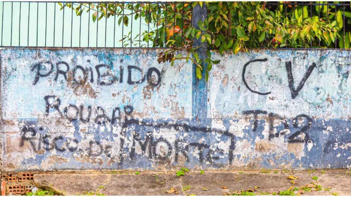 25% dos brasileiros vivem sob jugo de facções-Muro desgastado com pichações em tinta preta, incluindo mensagens de proibição e siglas de facção criminosa.