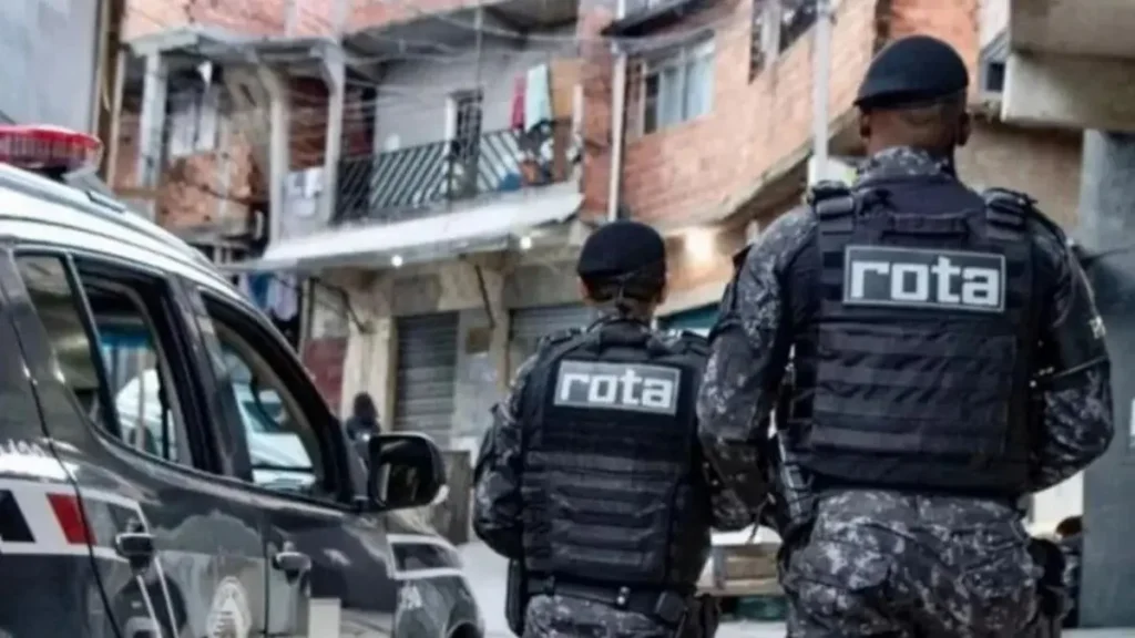A imagem mostra dois policiais da ROTA, unidade de elite da Polícia Militar de São Paulo, patrulhando uma comunidade com casas simples e ruas estreitas, ao lado de uma viatura.