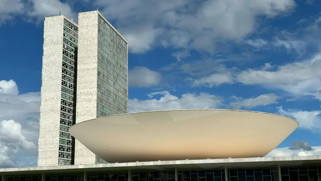 Vista do Congresso Nacional em Brasília, com as torres gêmeas e a cúpula branca em destaque sob o céu azul com nuvens