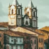 A imagem mostra construções históricas com fachadas antigas e coloridas em primeiro plano, e ao fundo duas torres de uma igreja colonial, com telhados de telha cerâmica e arquitetura barroca.