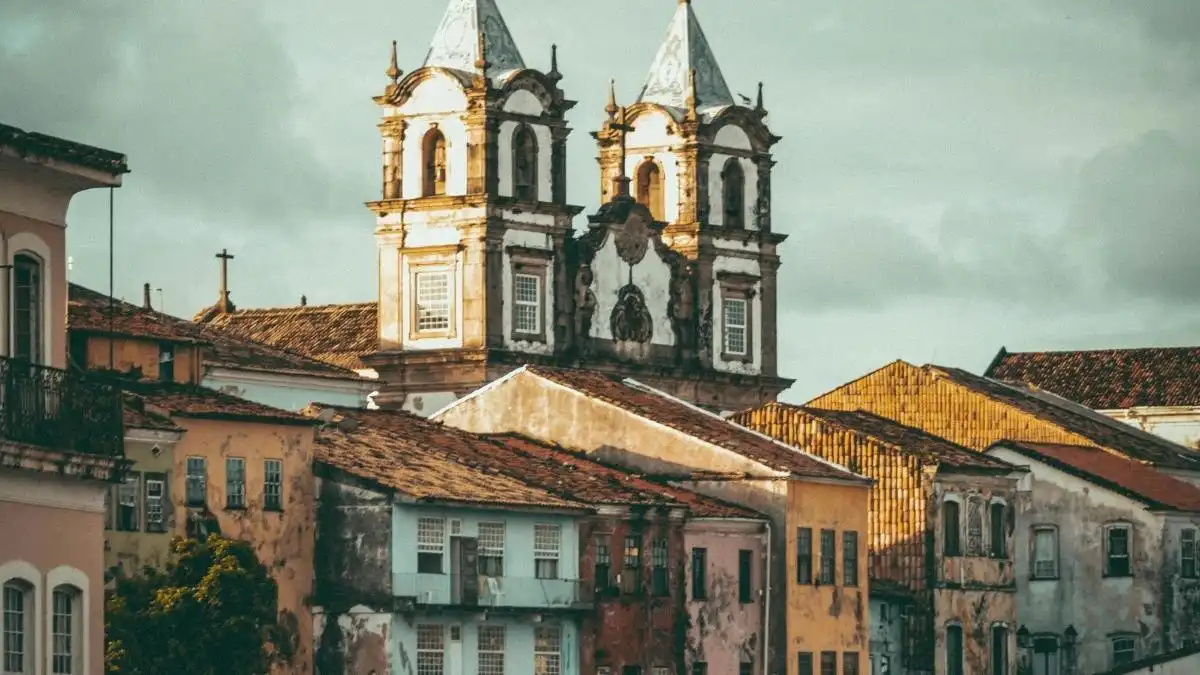 A imagem mostra construções históricas com fachadas antigas e coloridas em primeiro plano, e ao fundo duas torres de uma igreja colonial, com telhados de telha cerâmica e arquitetura barroca.