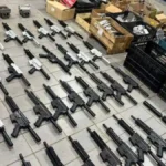 A imagem mostra dezenas de fuzis enfileirados no chão, em diferentes estágios de montagem, com cores preta e prateada. Ao fundo, há caixas e caixotes cheios de peças, evidenciando um arsenal clandestino desmontado em operação policial.