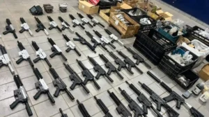 A imagem mostra dezenas de fuzis enfileirados no chão, em diferentes estágios de montagem, com cores preta e prateada. Ao fundo, há caixas e caixotes cheios de peças, evidenciando um arsenal clandestino desmontado em operação policial.