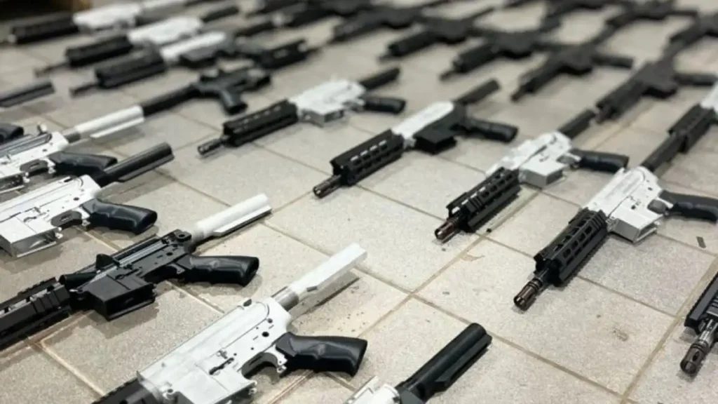 A imagem mostra várias armas longas, semelhantes a fuzis, dispostas no chão em fileiras organizadas. Algumas peças estão em tons metálicos prateados e outras em preto, indicando modelos em diferentes estágios de montagem ou finalização