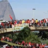 Bombeiros-A imagem mostra uma manifestação com várias pessoas vestindo roupas vermelhas reunidas sobre uma passarela e no gramado abaixo. Elas exibem bandeiras e faixas de protesto. Ao fundo, aparece o Pão de Açúcar, no Rio de Janeiro, como paisagem marcante.