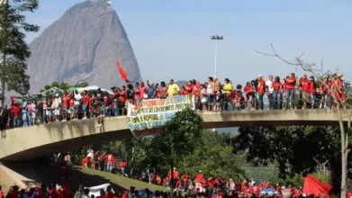 Bombeiros-A imagem mostra uma manifestação com várias pessoas vestindo roupas vermelhas reunidas sobre uma passarela e no gramado abaixo. Elas exibem bandeiras e faixas de protesto. Ao fundo, aparece o Pão de Açúcar, no Rio de Janeiro, como paisagem marcante.