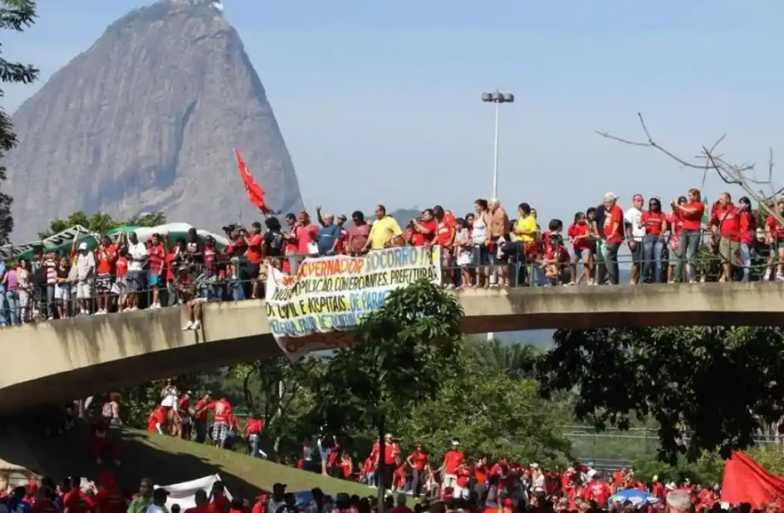 Bombeiros-A imagem mostra uma manifestação com várias pessoas vestindo roupas vermelhas reunidas sobre uma passarela e no gramado abaixo. Elas exibem bandeiras e faixas de protesto. Ao fundo, aparece o Pão de Açúcar, no Rio de Janeiro, como paisagem marcante.
