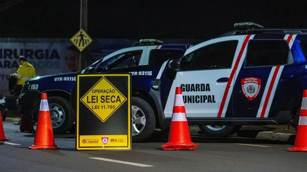 A imagem mostra uma operação Lei Seca realizada pela Guarda Municipal de Londrina. Em primeiro plano há um placar amarelo com os dizeres “Operação Lei Seca – Lei 11.705.