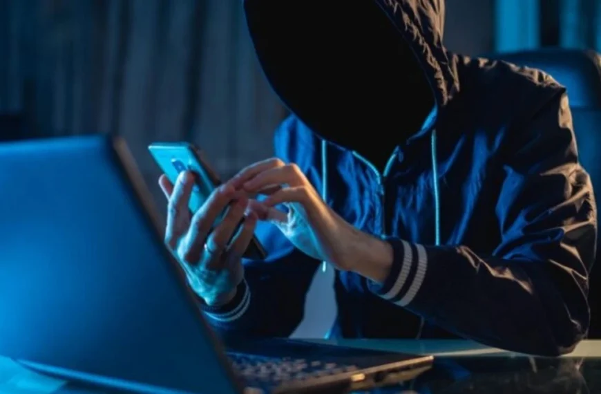 Sofreu golpe digital? Ele foi investigado? Pessoa com capuz escuro cobre o rosto enquanto manuseia um celular diante de um notebook, em ambiente de pouca luz, representando atividade clandestina ligada a crimes virtuais ou golpes digitais.