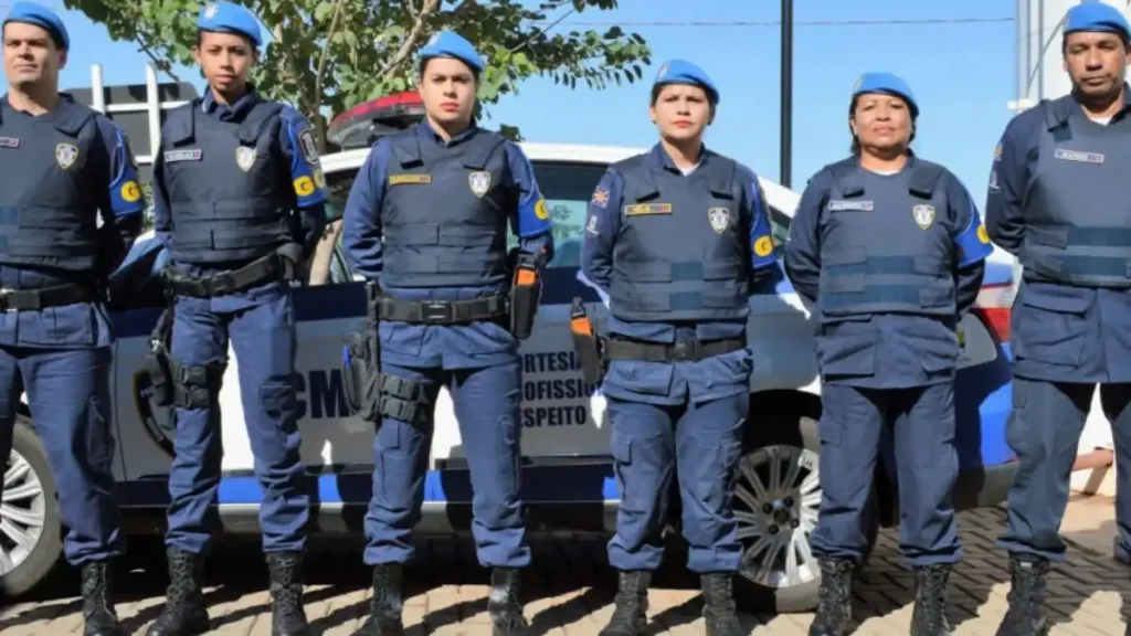 Seis agentes da Guarda Civil Metropolitana de Campo Grande (MS) posam uniformizados, com boinas azuis e coletes táticos, perfilados em frente a uma viatura oficial da corporação estacionada.