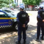 GCM de Campo Grande recebe investimentos-Dois agentes da Guarda Civil Metropolitana de Campo Grande (MS) aparecem de costas, uniformizados e de colete, ao lado de uma viatura oficial azul e amarela estacionada em via urbana movimentada.