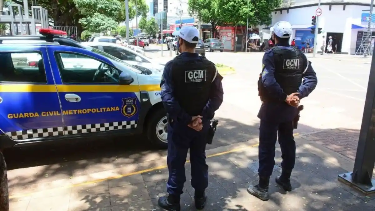 GCM de Campo Grande recebe investimentos-Dois agentes da Guarda Civil Metropolitana de Campo Grande (MS) aparecem de costas, uniformizados e de colete, ao lado de uma viatura oficial azul e amarela estacionada em via urbana movimentada.