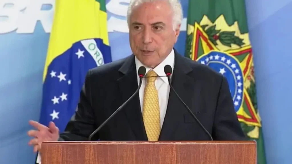 A imagem mostra um homem de terno escuro, camisa branca e gravata amarela, falando em um púlpito com dois microfones. Ao fundo, aparecem bandeiras do Brasil e símbolos oficiais, indicando um discurso em ambiente governamental.