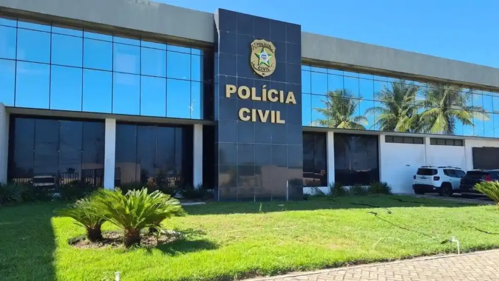 A imagem mostra a fachada de um prédio da Polícia Civil. O edifício tem paredes espelhadas, com destaque para uma estrutura central de cor escura onde estão o brasão e a inscrição “POLÍCIA CIVIL” em letras douradas. Em frente, há área gramada e alguns veículos estacionados.

