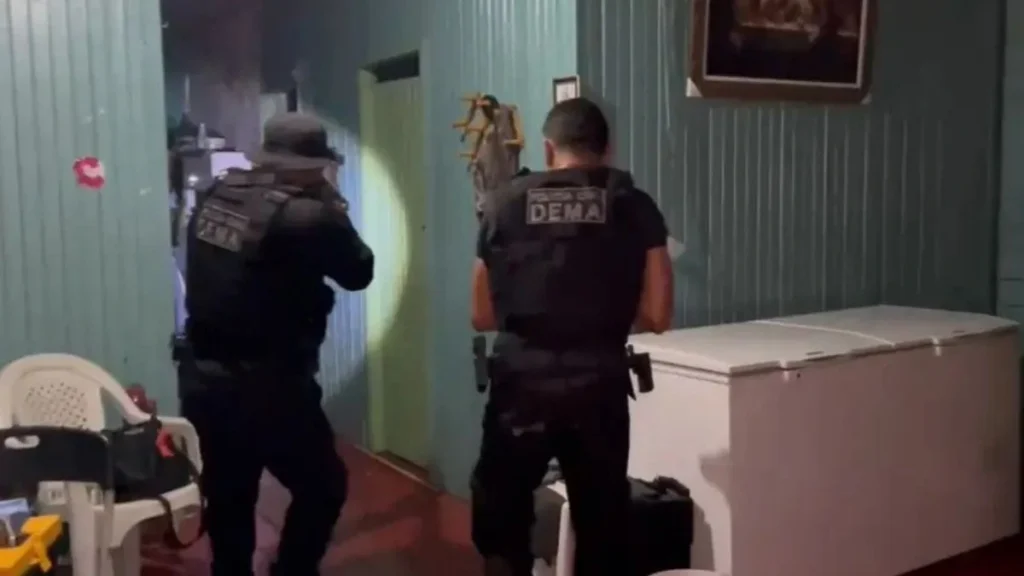 Dois policiais armados, usando coletes táticos com a inscrição “DEMA”, avançam por um corredor estreito de madeira durante operação noturna; um deles ilumina o ambiente com lanterna.
