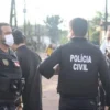 300 mil mandados em aberto-Três agentes da Polícia Civil conversam em uma rua de bairro residencial durante operação, todos usando coletes e máscaras de proteção. Ao fundo, há casas simples e viaturas estacionadas.