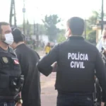 300 mil mandados em aberto-Três agentes da Polícia Civil conversam em uma rua de bairro residencial durante operação, todos usando coletes e máscaras de proteção. Ao fundo, há casas simples e viaturas estacionadas.