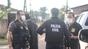 300 mil mandados em aberto-Três agentes da Polícia Civil conversam em uma rua de bairro residencial durante operação, todos usando coletes e máscaras de proteção. Ao fundo, há casas simples e viaturas estacionadas.