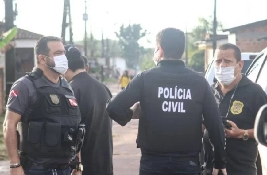 300 mil mandados em aberto-Três agentes da Polícia Civil conversam em uma rua de bairro residencial durante operação, todos usando coletes e máscaras de proteção. Ao fundo, há casas simples e viaturas estacionadas.
