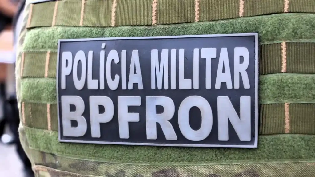 **Texto alternativo resumido:**
Close do colete tático de um policial com o brasão “Polícia Militar BPFron”, em referência ao Batalhão de Fronteira do Paraná.
