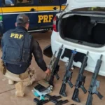 O corredor livre das armas ilegais- Policial rodoviário federal ajoelhado ao lado de um carro branco com o porta-malas aberto, onde estão apreendidos vários fuzis e carregadores; ao fundo, viatura da PRF estacionada.