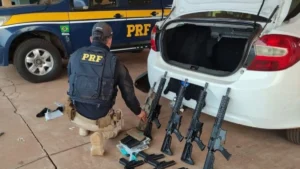 O corredor livre das armas ilegais- Policial rodoviário federal ajoelhado ao lado de um carro branco com o porta-malas aberto, onde estão apreendidos vários fuzis e carregadores; ao fundo, viatura da PRF estacionada.