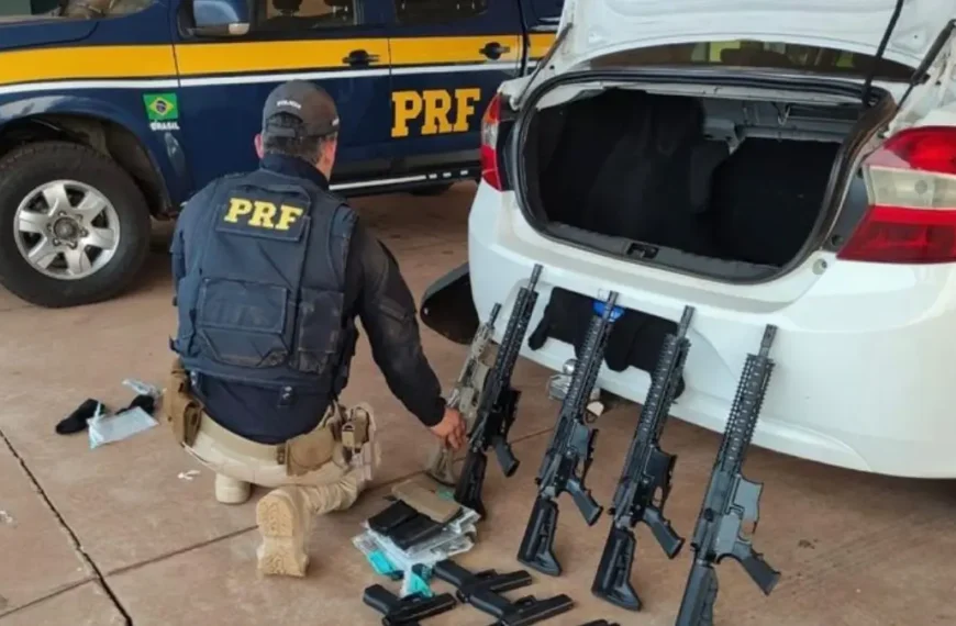 O corredor livre das armas ilegais- Policial rodoviário federal ajoelhado ao lado de um carro branco com o porta-malas aberto, onde estão apreendidos vários fuzis e carregadores; ao fundo, viatura da PRF estacionada.