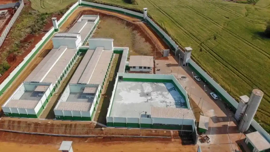 A imagem mostra uma visão aérea de um complexo penitenciário, com pavilhões, pátio murado e torres de vigilância, cercado por muros altos e localizado em área rural.