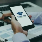 Pessoa utiliza smartphone exibindo o aplicativo da Receita Federal enquanto consulta documentos e realiza cálculos em uma mesa de trabalho.