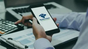 Pessoa utiliza smartphone exibindo o aplicativo da Receita Federal enquanto consulta documentos e realiza cálculos em uma mesa de trabalho.