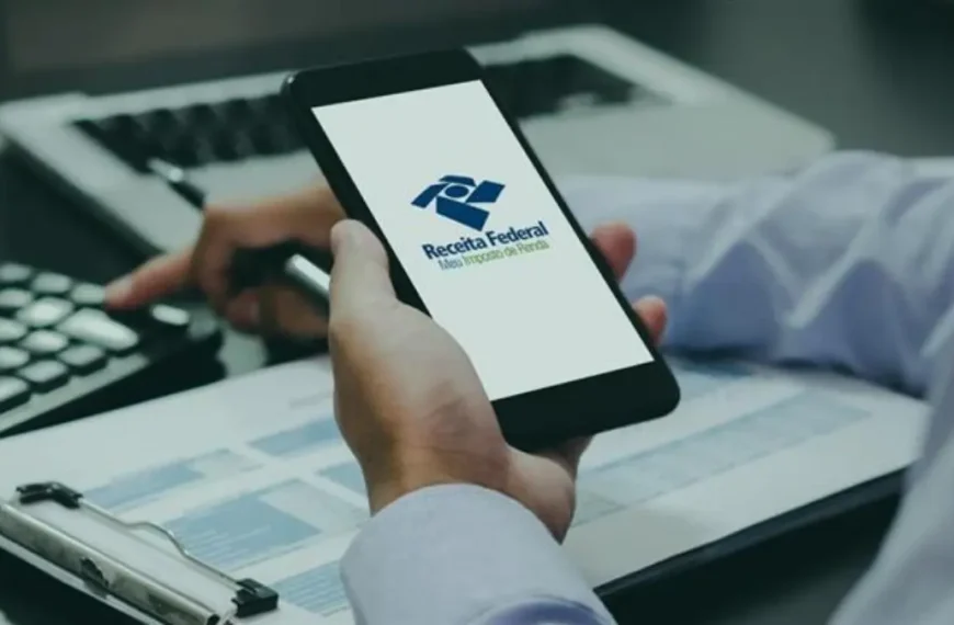 Pessoa utiliza smartphone exibindo o aplicativo da Receita Federal enquanto consulta documentos e realiza cálculos em uma mesa de trabalho.