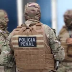 Três policiais penais com uniformes camuflados e equipamentos de proteção conversam em área interna de unidade prisional. Um deles está de costas, com identificação “Polícia Penal” no colete.