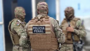 Três policiais penais com uniformes camuflados e equipamentos de proteção conversam em área interna de unidade prisional. Um deles está de costas, com identificação “Polícia Penal” no colete.